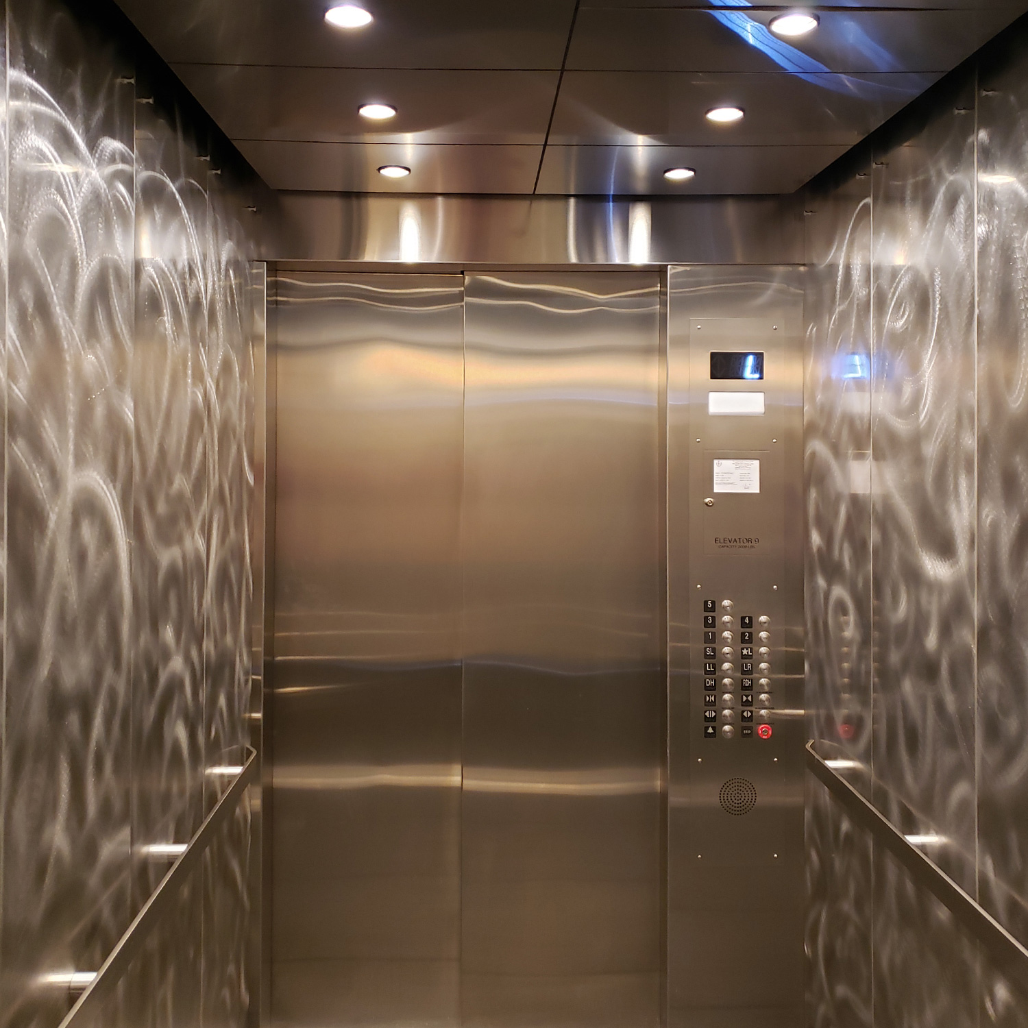 Patterned stainless steel elevator wall panels beneath warm recessed lighting